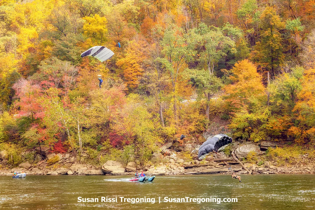 A parachutist descends toward a riverbank backed by dense autumn foliage, while a second canopy drifts low over the water preparing to splash down, with a couple of rescue boats nearby ready to assist jumpers making water landings.