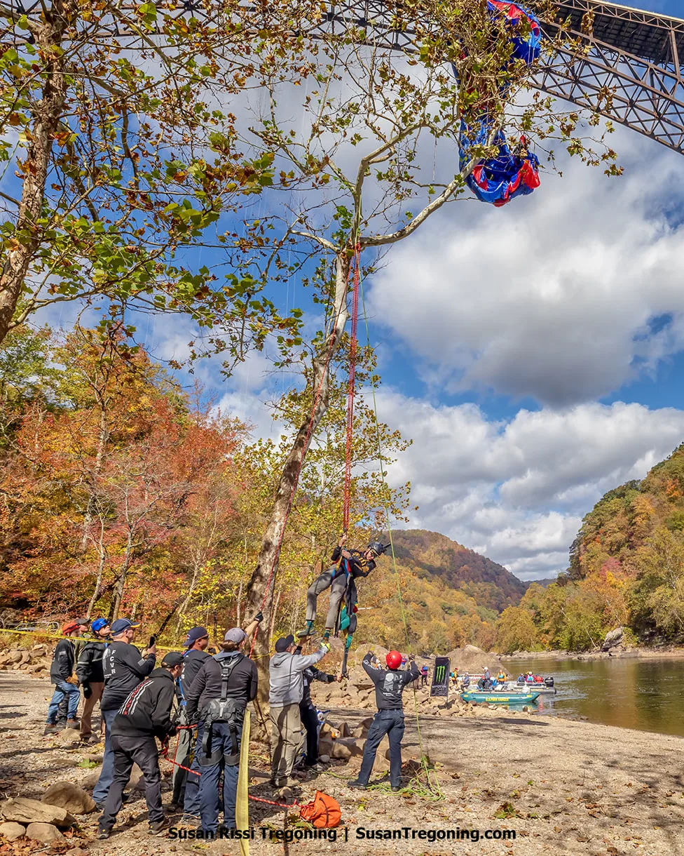 A BASE jumper is suspended in a harness as the Bridge Day rescue team works to lower him safely from a tree, with ropes, crew members, and autumn foliage visible along the riverbank below a steel bridge.