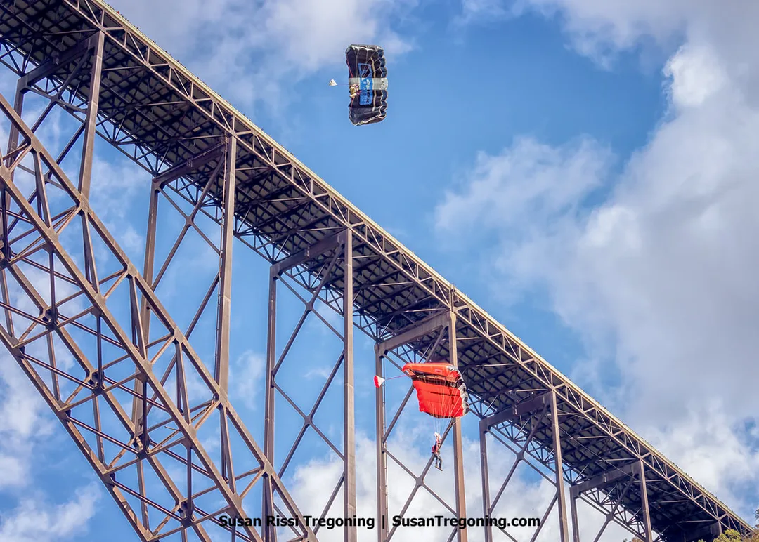 Two parachutists descend beside a large steel arch bridge, one under a black canopy and the other under a red canopy, set against a bright blue sky with scattered clouds and the bridge’s truss structure visible behind them.