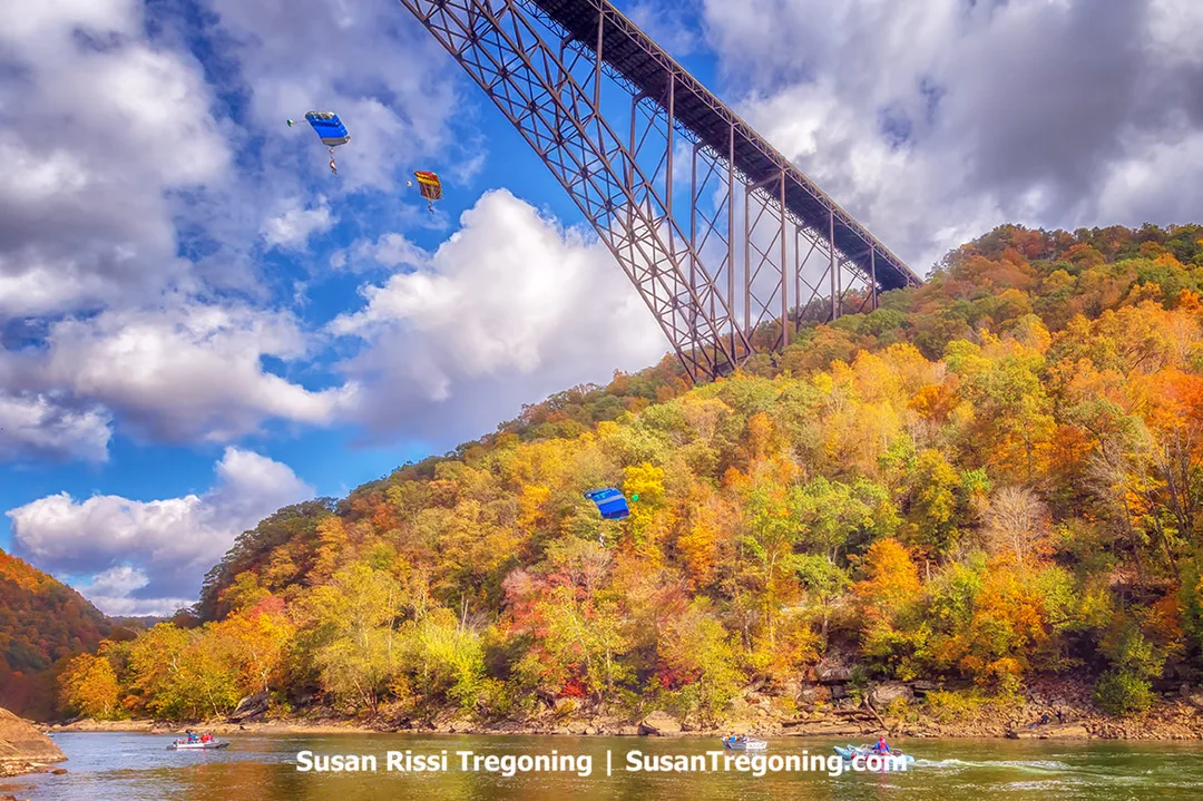 Parachutists with brightly colored canopies descend near a large steel arch bridge, drifting above a river that reflects the surrounding autumn foliage in the valley below.