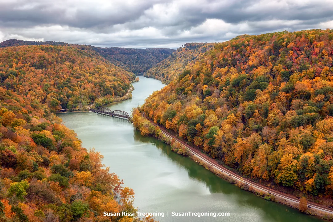 Aerial view of the New River Gorge at Hawks Nest State Park in West Virginia, showing a steel truss bridge spanning the river between forested hillsides with autumn foliage under an overcast sky.