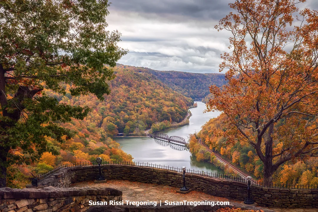 Stone terrace overlook with a flagstone platform, stacked stone walls, and a mounted binocular viewer, looking out over a winding river and forested hills with autumn foliage under an overcast sky.