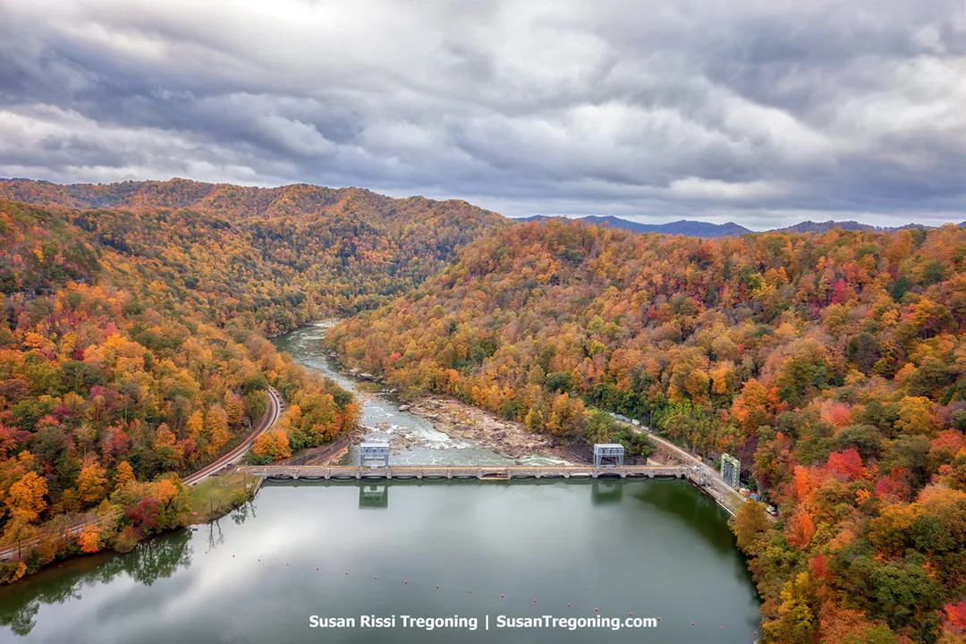 Aerial view of Hawks Nest Dam spanning the river, surrounded by forested hills with peak autumn foliage under an overcast sky.