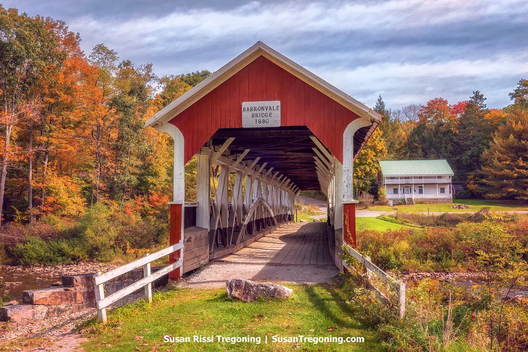 A red covered bridge with white trim spans a small creek, surrounded by autumn trees in shades of yellow, orange, and green. A sign above the entrance reads Barronvale Bridge 1880. The bridge’s two spans are different lengths, creating Burr Arches of varying heights within the structure. A white house and grassy area appear in the background.