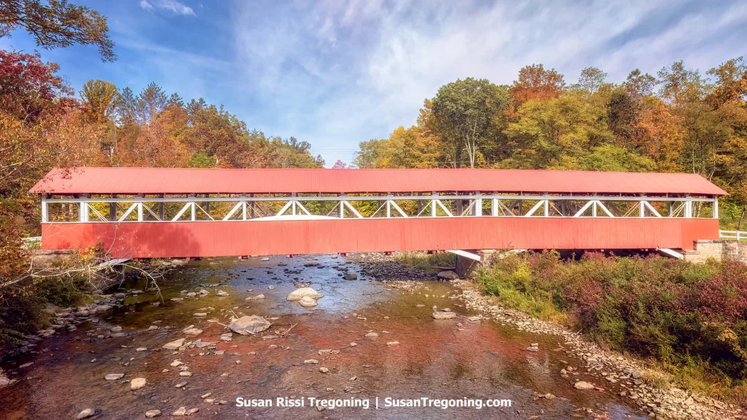 A long red covered bridge with white trim spans a shallow rocky river, surrounded by autumn trees in shades of green, yellow, orange, and red. The bridge rests on stone abutments and features open side panels with visible trusswork. It is the longest covered bridge in Somerset County.