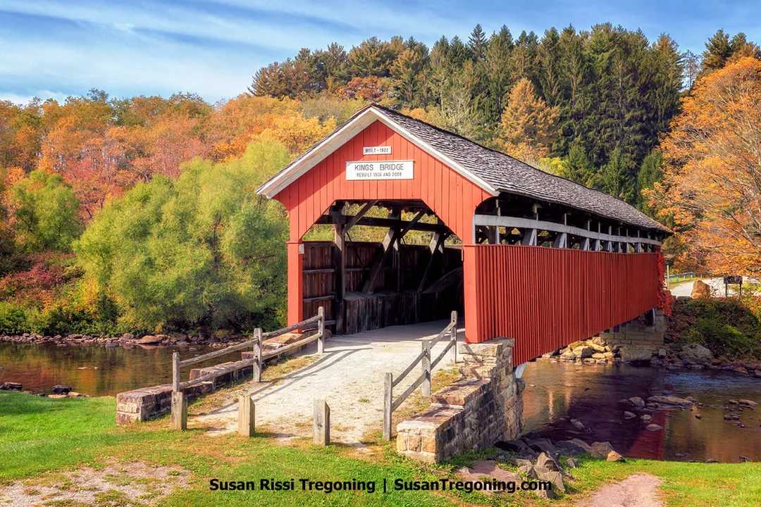 A red covered bridge with a shingled roof spans Laurel Hill Creek, surrounded by autumn trees in shades of yellow, orange, and orange‑brown. The bridge rests on stone abutments above flowing water and measures 127 feet in length. 