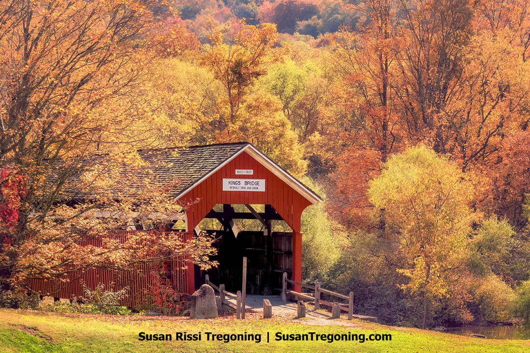 A red covered bridge with a gabled roof spans a small creek, surrounded by autumn trees in shades of orange, yellow, and green. A sign above the entrance reads Kings Bridge with construction and rebuilding dates of 1861, 1906, and 2008. Wooden railings lead toward the bridge, and fallen leaves cover the ground.