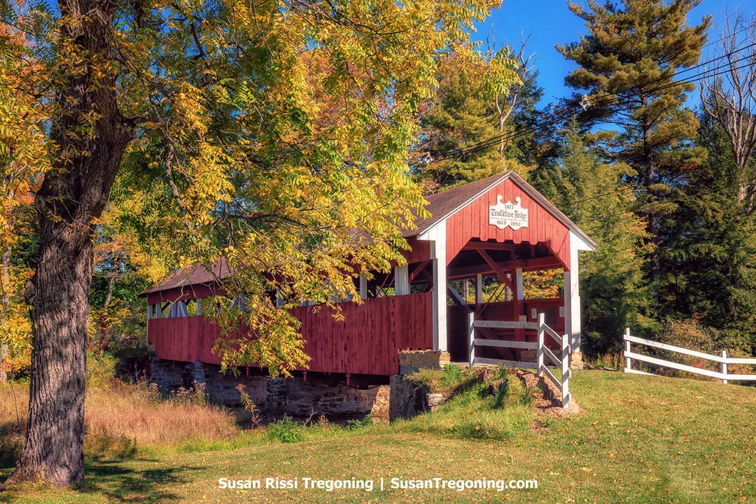 A red covered bridge with white trim and a gabled roof stands above a small stream, surrounded by trees with yellow and orange autumn foliage. A sign on the front of the bridge reads Cristina’s Bridge Built 1998. A white wooden fence lines the approach, and the bridge rests on stone foundations.