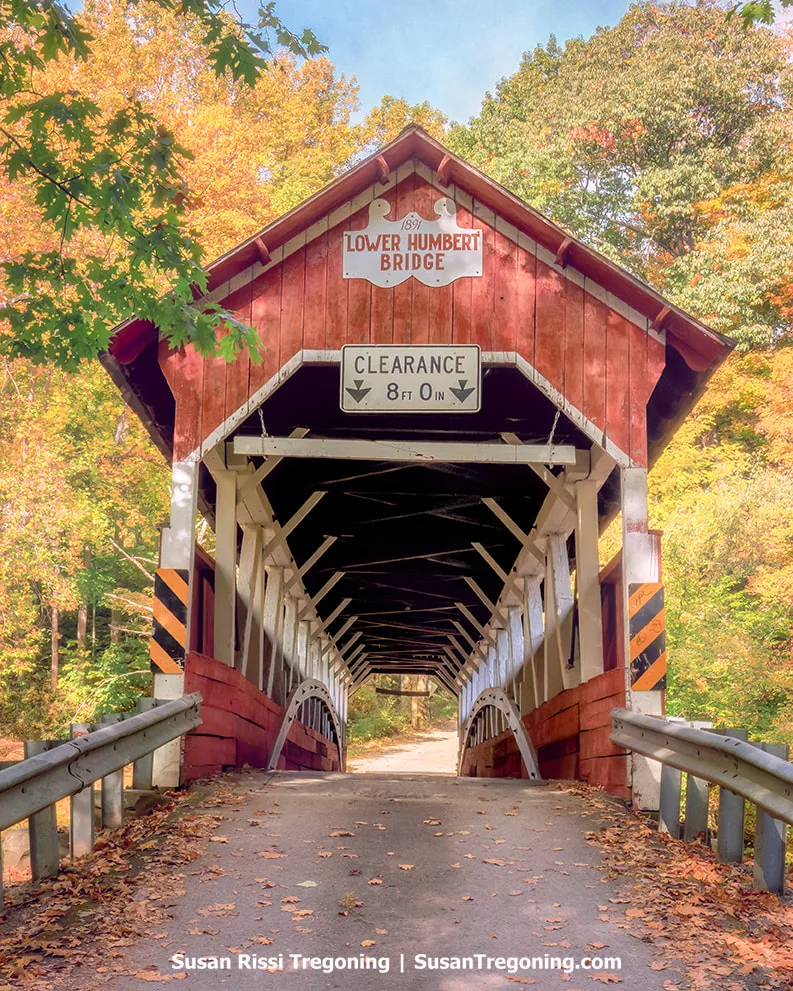   
A red covered bridge with white trim stands at the entrance to a forested roadway lined with autumn foliage in shades of orange, yellow, and green. A sign above the portal reads 1891 Lower Humbert Bridge, with a clearance sign below it. The wooden interior truss structure is visible through the opening. 