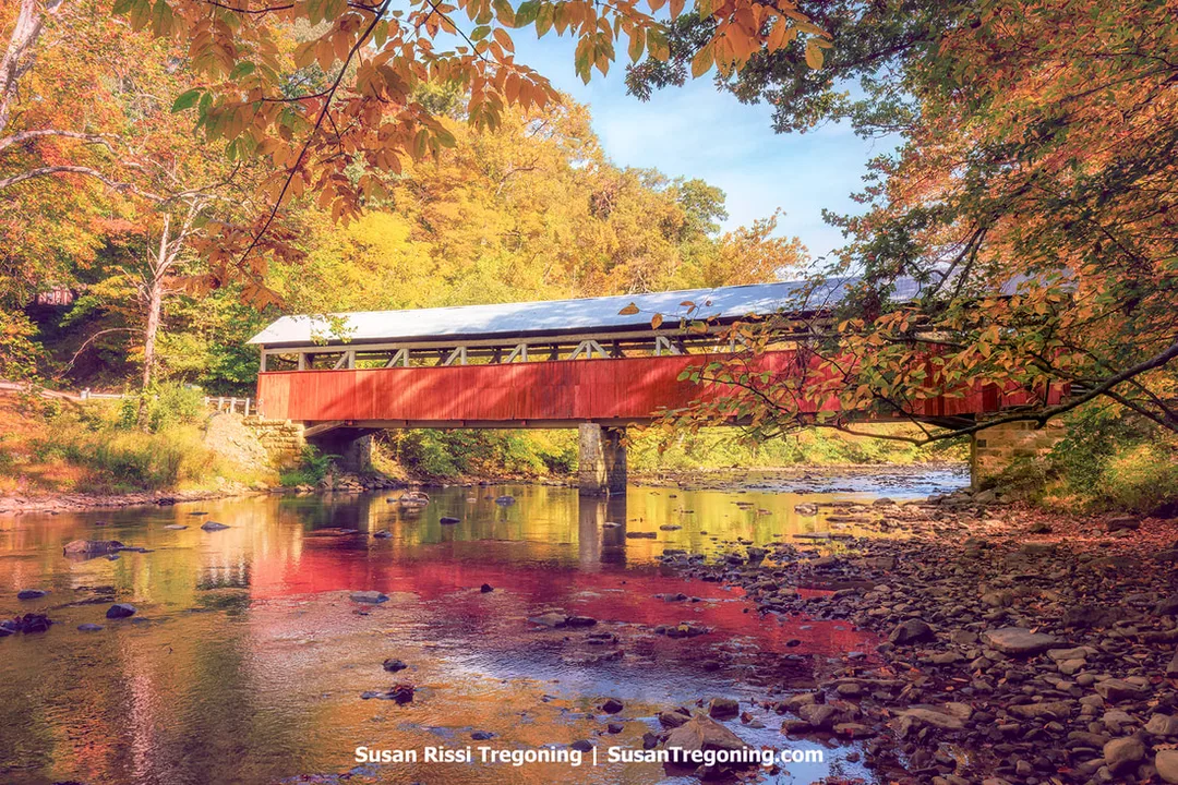 A red covered bridge with white trim spans Laurel Hill Creek, viewed in profile from the creek bank. The bridge rests on stone abutments above shallow flowing water, with autumn trees in shades of yellow, orange, and green surrounding the scene. The upper side panels of the bridge include open window slats beneath the roofline.