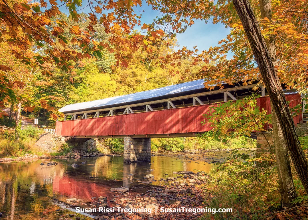 A red wooden covered bridge spans a calm stream surrounded by autumn trees in shades of orange, yellow, and green. The bridge has a metal roof and rests on stone supports. The fall foliage and the bridge are reflected in the still water below. Warm sunlight highlights the seasonal colors.