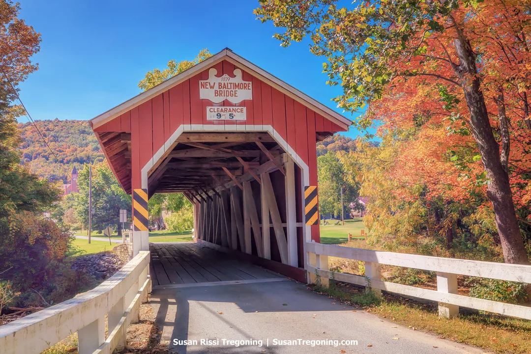 A red covered bridge with white trim spans a small creek, surrounded by autumn trees in shades of orange, yellow, and green. A sign above the entrance reads 1879 New Baltimore Bridge with a posted clearance of 9 feet 5 inches. Rolling hills and a clear sky appear in the background. The structure is a replica and the only covered bridge in Somerset County not listed on the National Register of Historic Places.