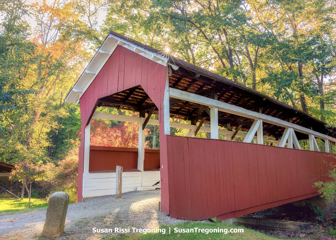 A red covered bridge with white trim stands at the end of a gravel path, surrounded by trees with green and early autumn leaves. Sunlight filters through the branches, illuminating the wooden beams inside the bridge and the foliage around it.