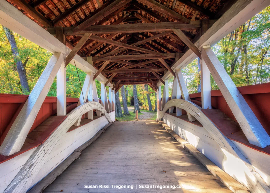 Interior view of a covered wooden bridge with exposed beams, a peaked roof, and curved side supports. The floor is made of wooden planks, and the railings have white trim with red panels. Trees with green foliage are visible through the openings at the far end of the bridge.