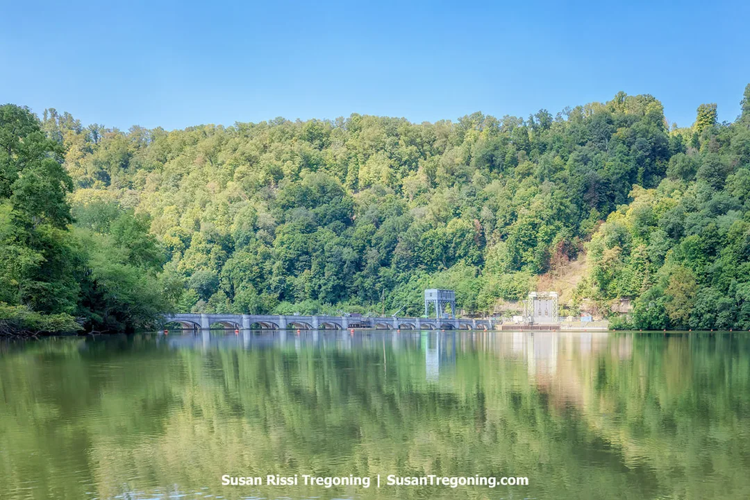 Hawks Nest Dam spanning the river, reflected in the calm backwater of Hawks Nest Lake, with forested green hills under a clear blue sky.