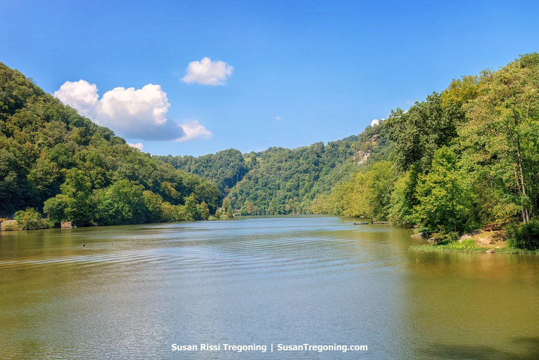 Calm water on Hawks Nest Lake in West Virginia, surrounded by densely forested green hills  of the New River Gorge under a blue sky with scattered white clouds.