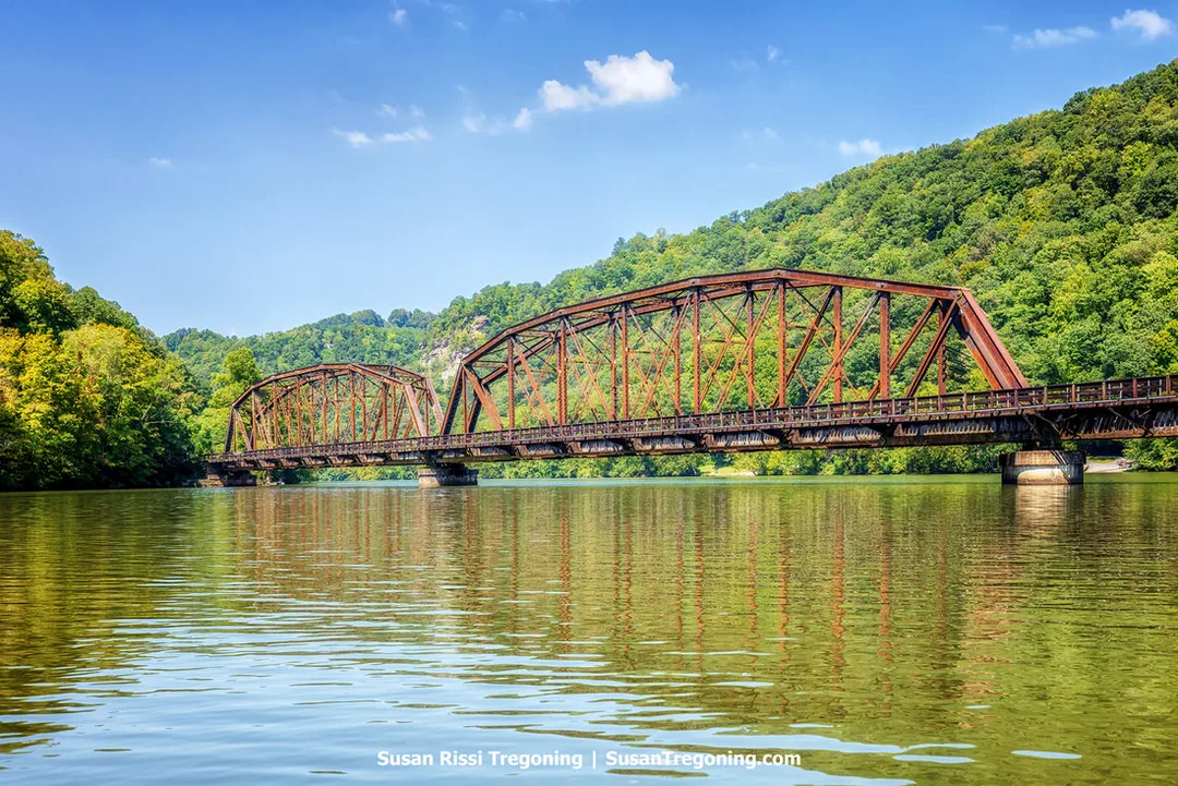 A rust‑colored steel truss bridge spans a calm river, surrounded by forested hills under a blue sky with scattered clouds.