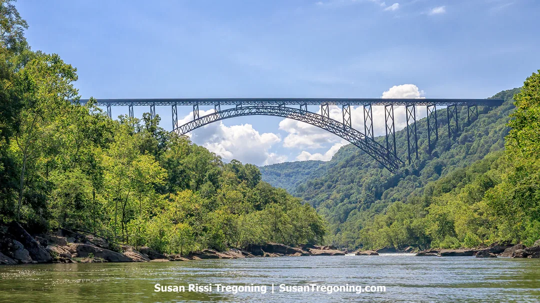 A large steel arch bridge spans the New River Gorge, rising high above the river and surrounded by forested hills. The view is seen from the New River Gorge Jet Boat tour area at Hawks Nest State Park, with blue sky and scattered clouds above the gorge.