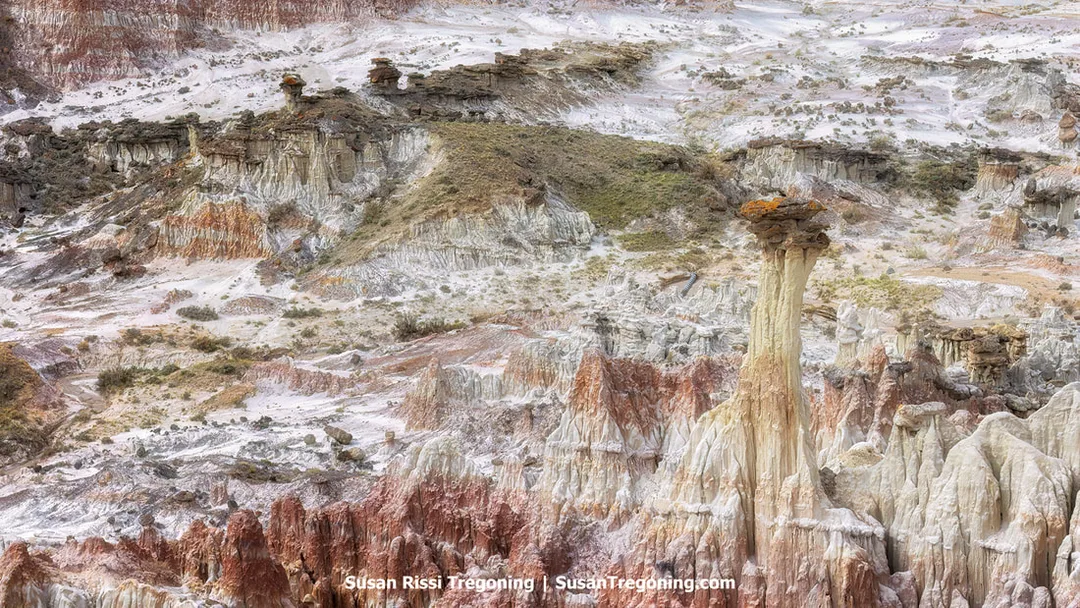 Colorful badlands formations with layered red, white, and tan rock rise in eroded cliffs and hoodoo-like shapes, with sparse vegetation scattered across the slopes.