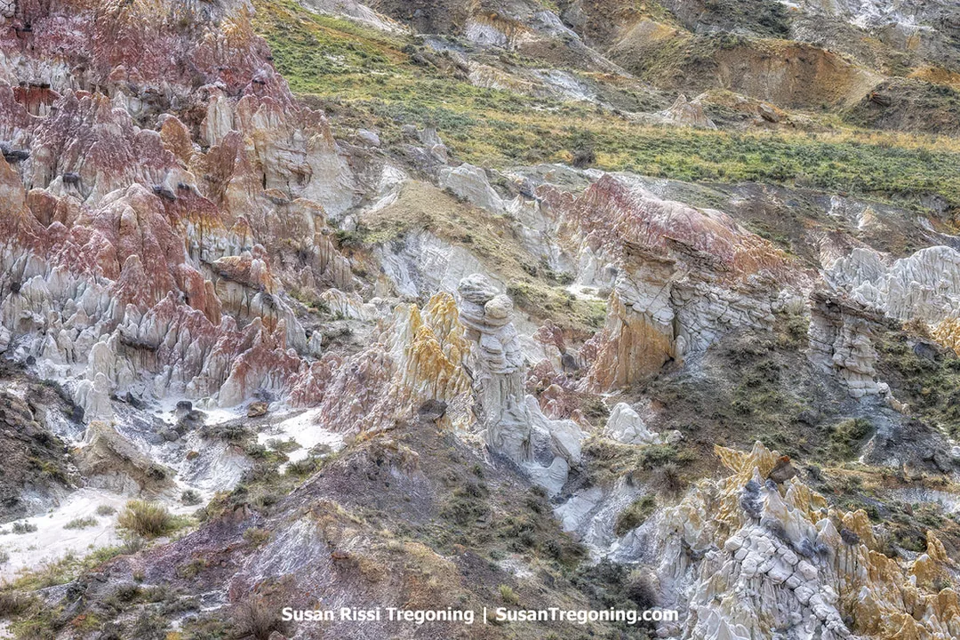 Colorful badlands formations with layered red, orange, yellow, white, and gray rock rise in eroded cliffs and hoodoo-like shapes, with sparse vegetation on the upper slopes.