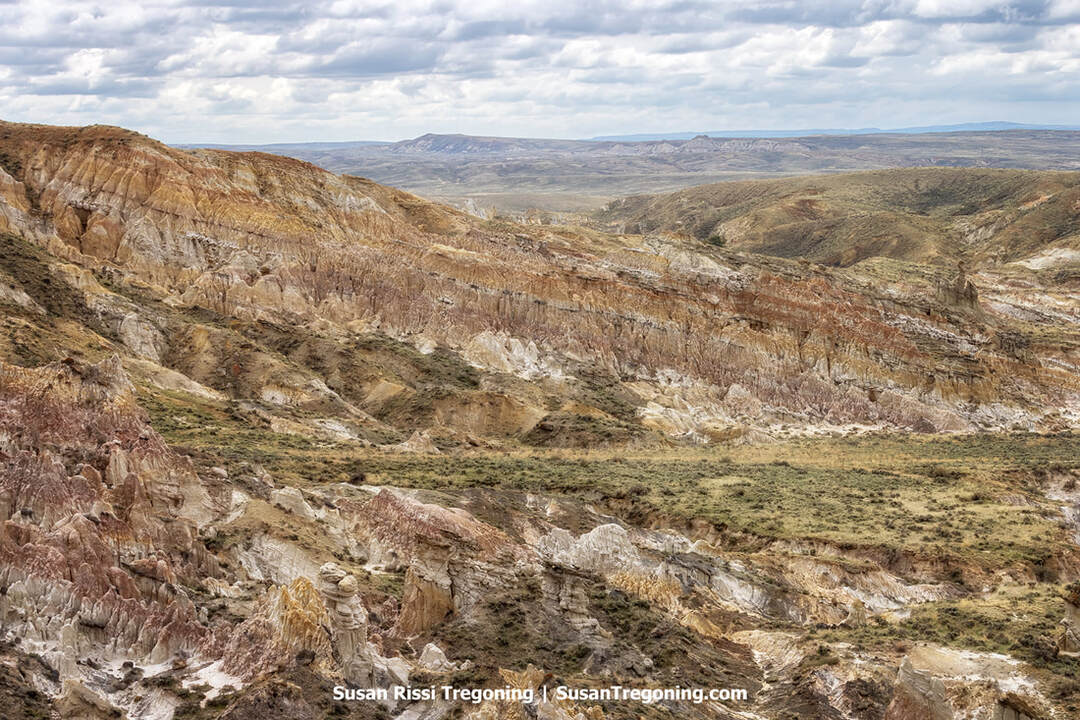 Eroded badlands formations with layered tan, brown, white, and reddish rock rise in rugged slopes and cliffs, with sparse vegetation and distant hills beneath an overcast sky.