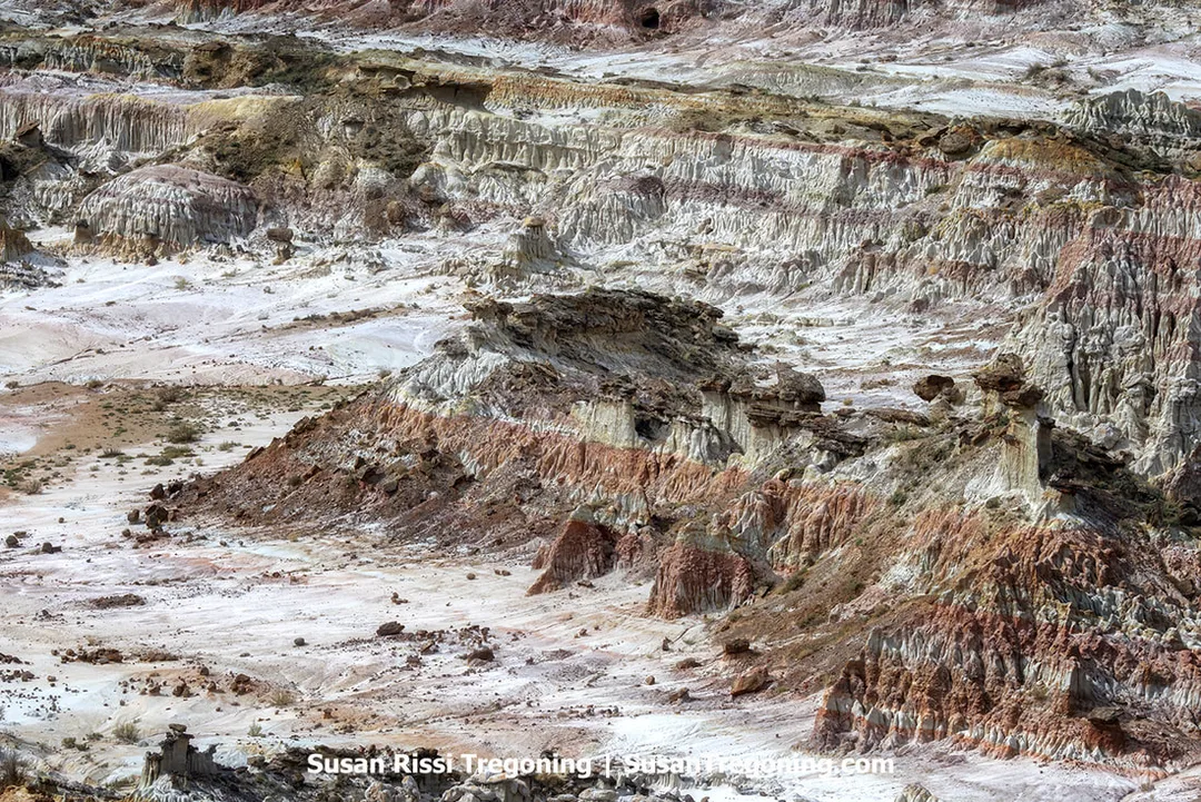 Layered badlands formations in shades of white, tan, red, and brown rise in eroded cliffs and ridges, with sparse vegetation on the valley floor and distant hills in the background.