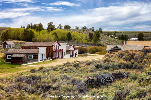 An old wooden wagon rests in sagebrush on a hillside overlooking South Pass City. Behind them are preserved nineteenth‑century wooden buildings set among rolling hills, trees, and open grassland beneath a partly cloudy sky.
