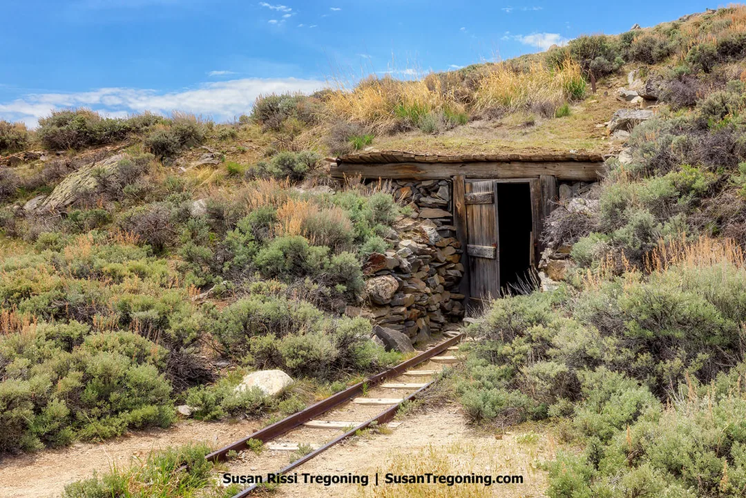 Historic mine entrance constructed of stacked stone and timber, set into a hillside with a partially open wooden door leading into a dark tunnel. Narrow‑gauge rails run from the doorway into surrounding sagebrush and grasses under a partly cloudy sky.