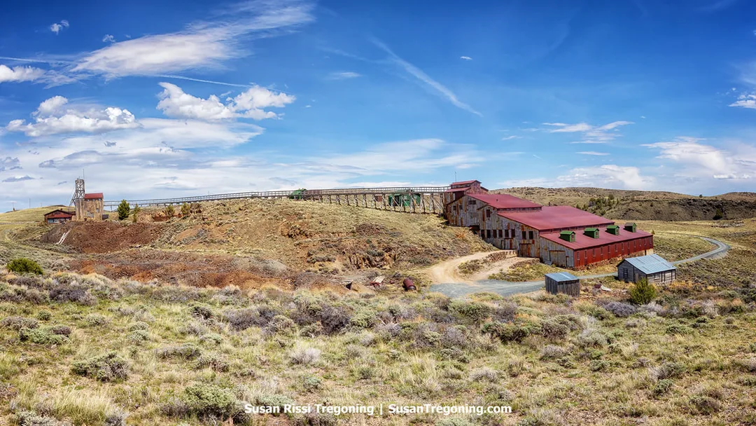 View of the Carissa Mine and Mill in South Pass City, with preserved wooden and metal buildings, trestles, and mining structures spread across open grassland. Rolling hills and a partly cloudy sky frame the historic gold‑mining site.
