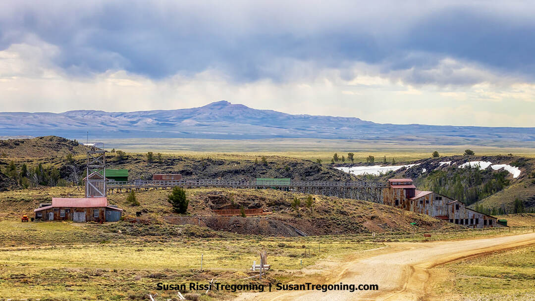 Wide view of the Carissa Mine and Mill at South Pass City, showing weathered wooden and metal buildings, a tall framework tower, and a long elevated trestle on open grassland. Dark clouds hang over the surrounding mountains.