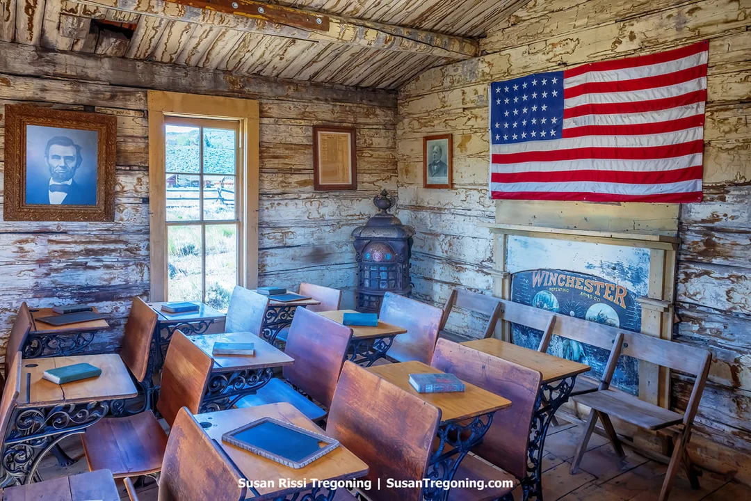 The interior of a one‑room schoolhouse with wooden desks arranged in rows, a potbelly stove at the back, framed pictures on the walls, and a 48‑star American flag hanging above a window.