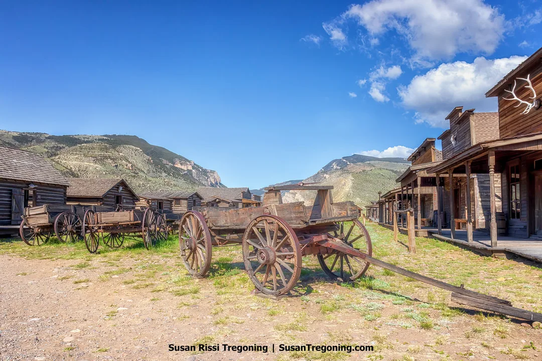 A row of wooden wagons with large spoked wheels positioned between rustic wooden buildings in a historic Old West town setting, with mountains and a partly cloudy sky in the background.
