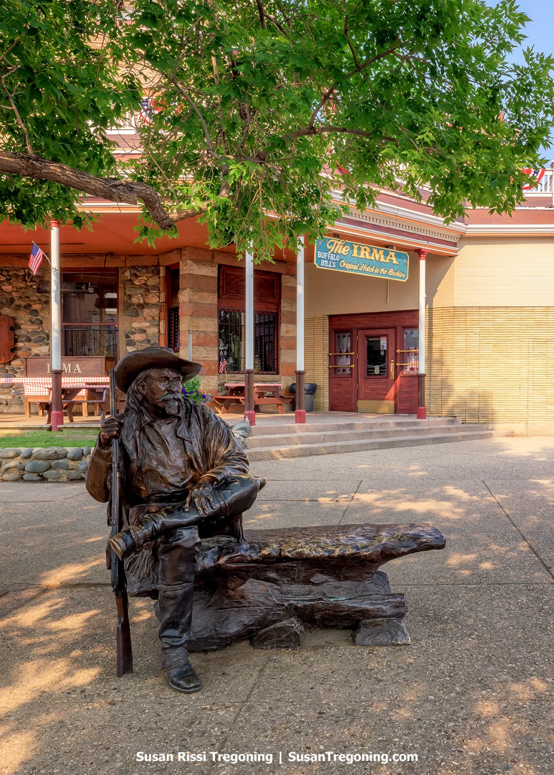 A bronze sculpture of Buffalo Bill Cody seated on a bench outside the Irma Hotel in Cody, Wyoming, with the hotel’s stone and brick façade and signage visible behind him.