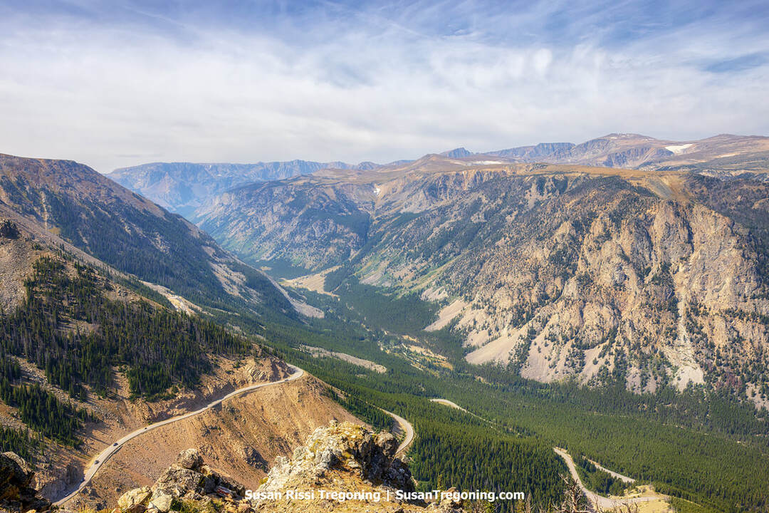 A high‑elevation view across steep rocky slopes and dense forested areas, with a winding road following the valley floor. Multiple layers of distant mountain ridges extend toward the horizon beneath a partly cloudy sky.