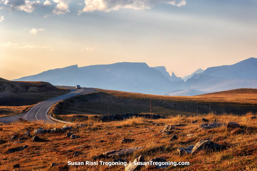A winding paved road curves through open grassy terrain toward distant hazy mountains. A vehicle travels along the road, with rocky slopes, scattered grass, and layered mountain ridges visible beneath a partly cloudy sky.
