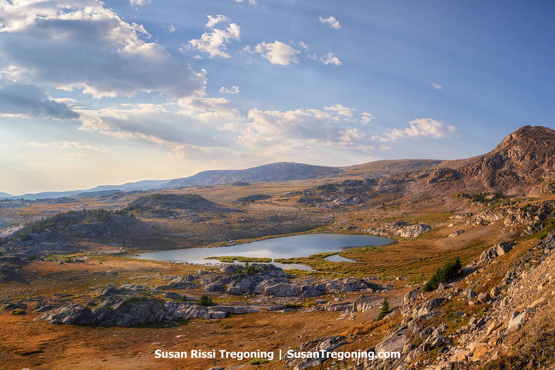 A high‑elevation alpine landscape with a small lake surrounded by rocky ground and open grassy areas. Rolling hills and distant mountain ridges extend toward the horizon beneath a partly cloudy sky.