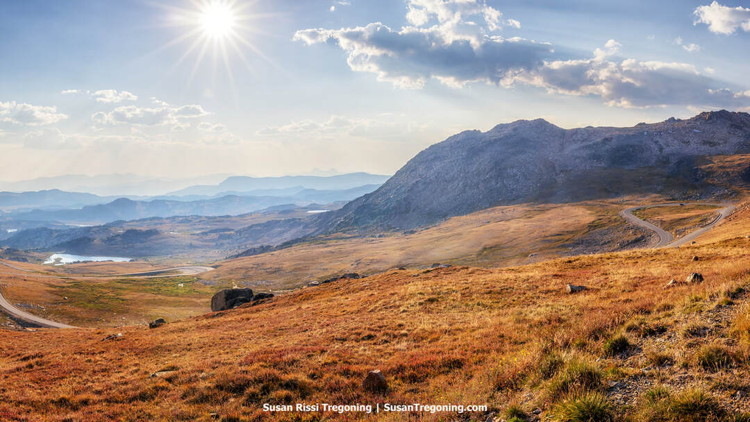 A high‑elevation landscape above the tree line with golden‑brown tundra, scattered rocks, and a winding paved road. A rugged mountain rises to the right, with distant hazy ridges and a small lake visible beneath a partly cloudy sky and bright sun.