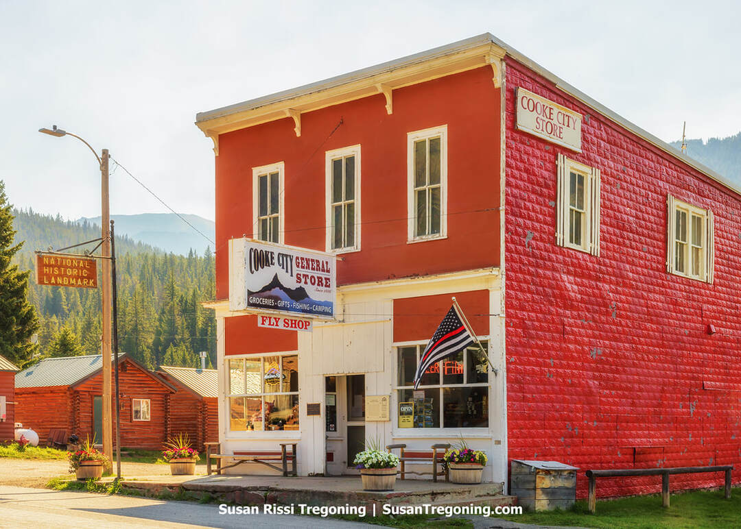 A two‑story red general store with white trim stands along a mountain‑town street, with signs for groceries, gifts, fishing, camping, and a fly shop. Flower planters sit near the entrance, an American flag hangs beside the door, and forested hills rise in the background.