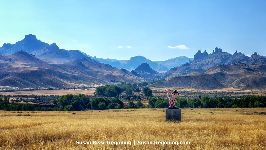A Big Boy statue stands on a concrete pedestal in an open field of golden grass. A few scattered buildings and trees sit farther back in the landscape, with rugged, jagged mountains rising sharply behind them under a clear blue sky.