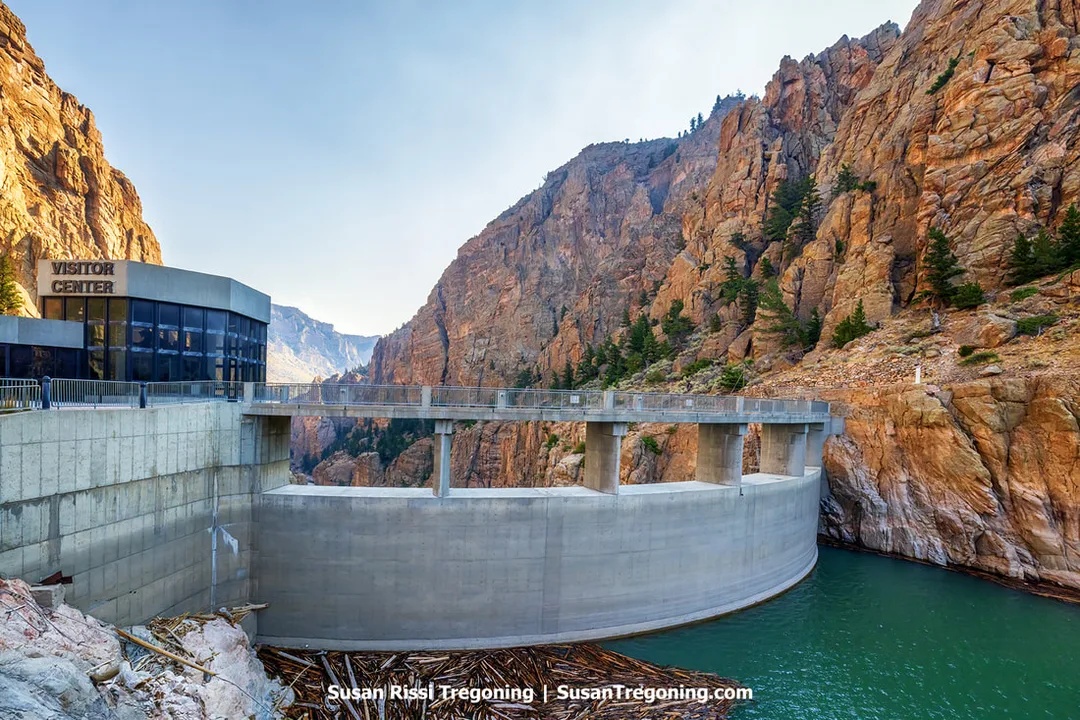 A large concrete dam spans a narrow rocky canyon, holding back greenish water. A modern visitor center sits on the left side of the dam, built against steep reddish‑brown canyon walls with scattered vegetation.