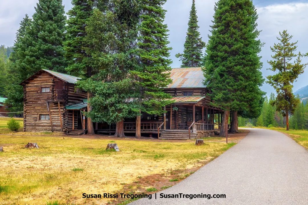 A rustic log lodge with a metal roof sits among tall evergreen trees, with a paved path leading through a grassy clearing toward the building. The cabin’s porch and large log construction are visible beneath a clear blue sky.