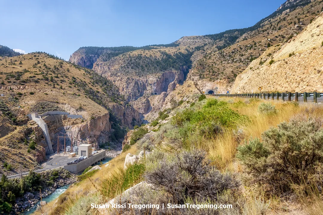 A high vantage view into a rocky mountain canyon showing a hydroelectric power plant built against the left canyon wall beside a narrow river. A paved road curves along the right side of the canyon and enters a short tunnel through the rock. The terrain is steep and rugged with sparse vegetation under a clear blue sky.
