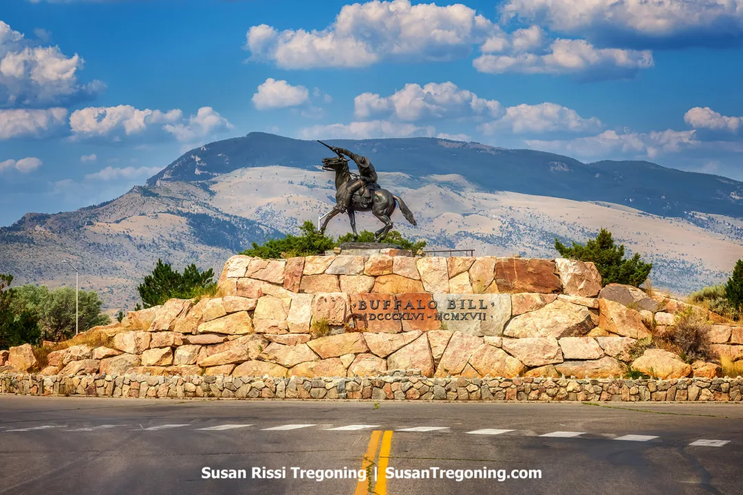 A bronze statue of a rider on a rearing horse set on a large stone base, with mountains and a partly cloudy sky in the background.