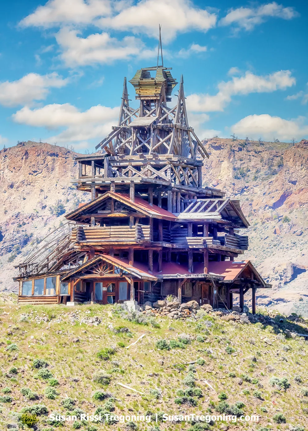   
A tall, multi‑story wooden structure with an irregular, log‑built design stands on a grassy hillside. The building features stacked balconies, exposed timber framing, and a tower‑like upper section. Rugged cliffs rise behind it under a bright blue sky with scattered clouds.
