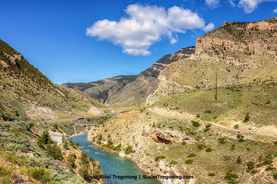 A wide view of a mountain canyon with the Shoshone River winding between steep rocky slopes and green hillsides, with a road and bridge visible in the distance under a blue sky with scattered clouds.