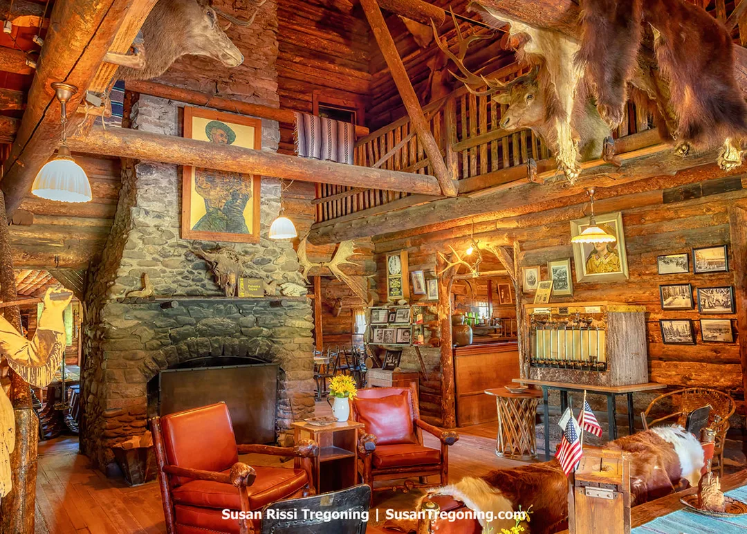 The interior of a rustic log lodge features a large stone fireplace with a painting above it, surrounded by log walls, wooden beams, and mounted animal heads. Leather chairs, a wooden table, framed photographs, and Western‑style décor fill the room, with a log staircase and balcony visible in the background under warm lighting.
