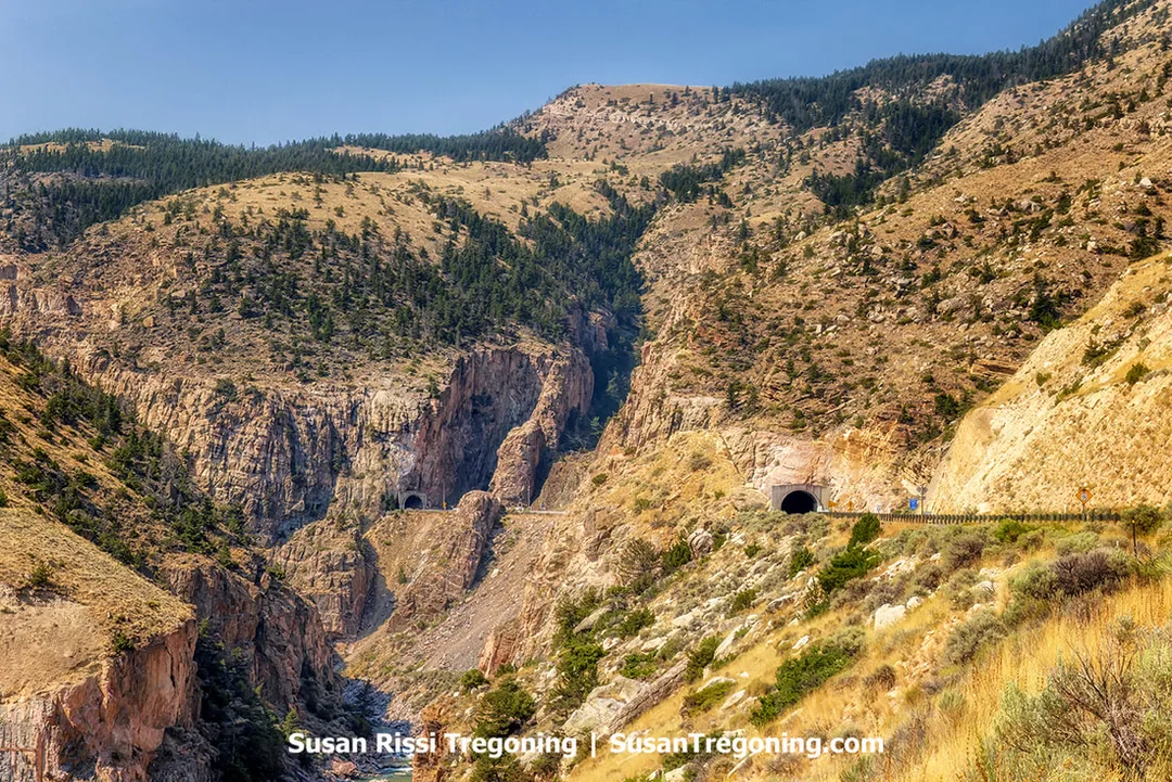 A view across a steep, rocky canyon where a paved road curves along the mountainside and passes through two separate tunnels carved into the cliffs. The terrain is dry and rugged with sparse shrubs and scattered trees on the slopes.