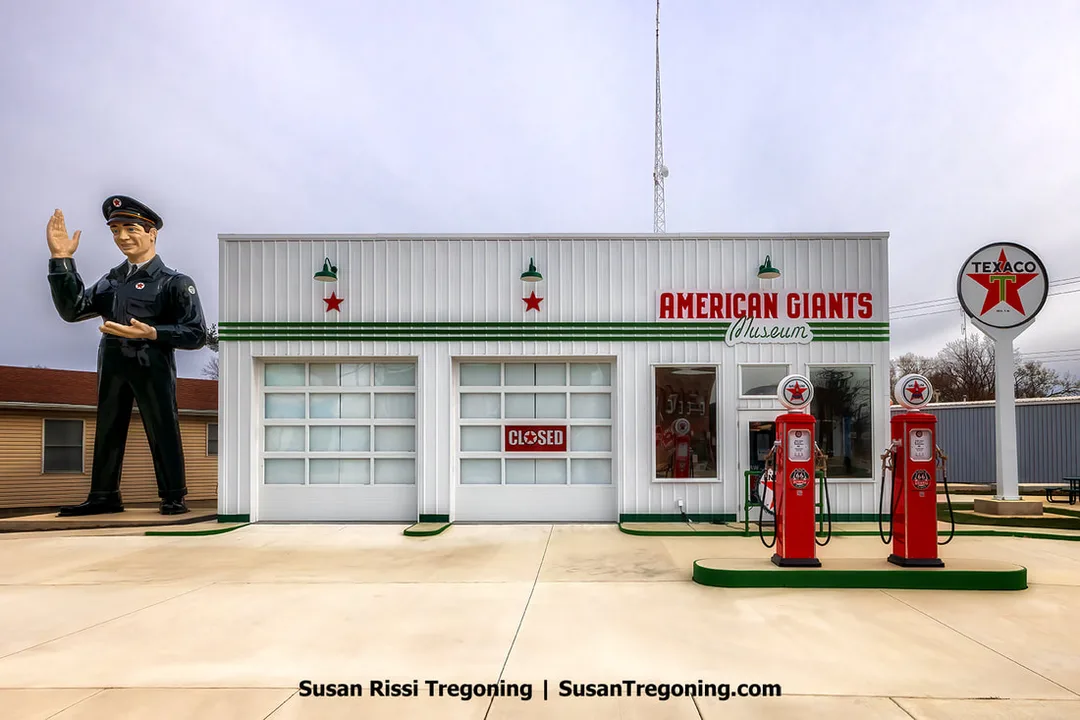 A rare Texas Big Friend Giant stands beside the American Giants Museum on the 1926 alignment of Route 66 in Atlanta, Illinois. The tall fiberglass figure is positioned near the Texaco Gas Station Museum building with surrounding streetscape elements visible.
