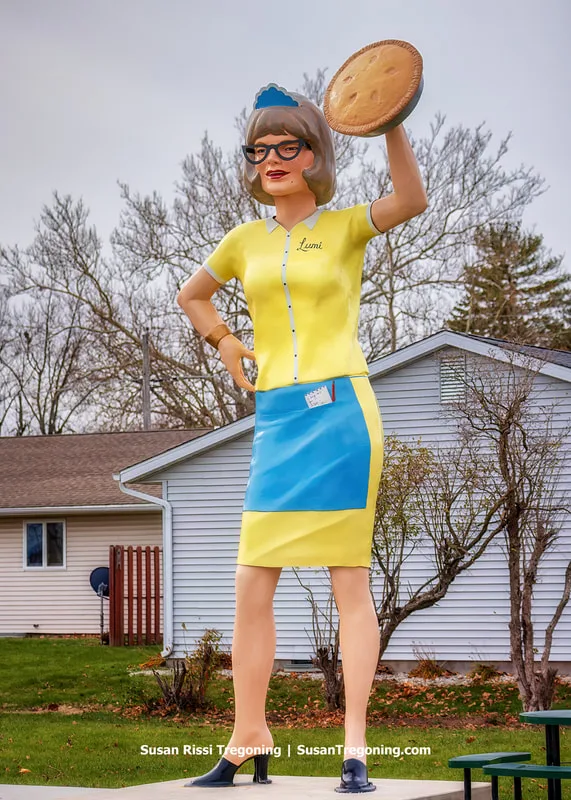 Lumi the Pie Lady, a 19‑foot‑tall fiberglass giant, stands outside the Country Aire Restaurant on Route 66 in Atlanta, Illinois. The figure wears a yellow dress, blue apron, cap, and cat‑eye glasses, with an order pad tucked into her pocket. She holds an apple pie raised in the air. Lumi is a variation of the Uniroyal Tire Gal design created by the International Fiberglass Company in the 1960s.