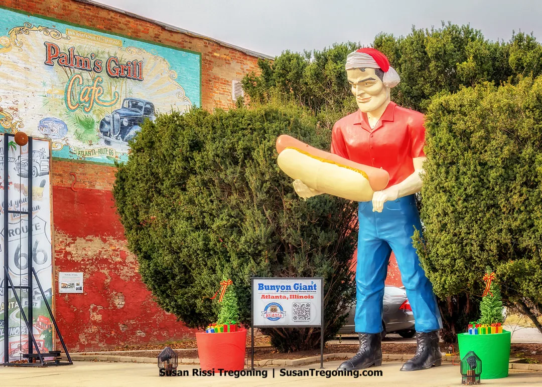 The Paul Bunyon Giant stands on Route 66 in Atlanta, Illinois, wearing a festive Christmas hat and holding an oversized hot dog. The figure is positioned between shrubs next to a building that features a Palms Grill Cafe mural on its side.